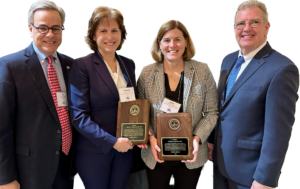 Shareholder Helen Casale in a tan jacket and black blouse next to Carolyn Zack wearing a blue suit and white blouse receiving wooden plaque awards. Gentlemen in bluesuits and white shirts are on either side.